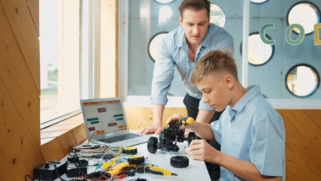 Caucasian teacher help student fixing car model and giving advise for programing system or coding prompt while highschool boy listen and use equipment to repair robot at STEM classroom. Edification.
