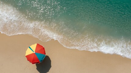 Obraz premium A single, colorful beach umbrella stands on a sandy beach, with a wave gently lapping at the shore. The turquoise water of the ocean stretches out to the horizon, creating a picturesque summer scene. 