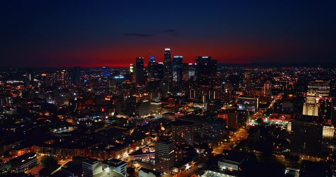 Skyscrapers of night Los Angeles with neon illumination on tops. Red sky above the metropolis. Drone flying over the city with bright lights.