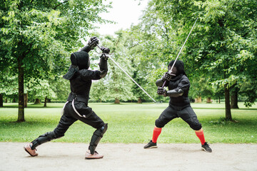 Two fencers dressed in black protective uniforms, helmets with face masks are fencing in city park...