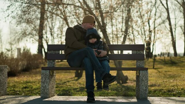 Father and son sitting on a bench in a park, the father rests his head gently on his son's head as he holds him close with leg crossed, with tress in the background