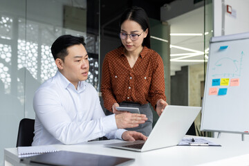 Asian business professionals engaged in collaborative work in modern office setting. Man and woman discussing project details, using laptop and phone. Teamwork technology in professional environment
