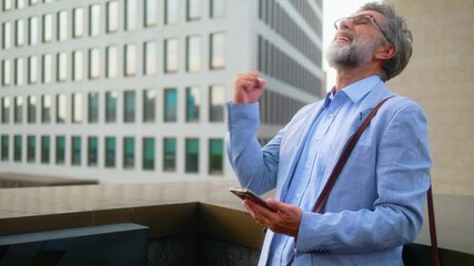 Elderly businessman reading mail or sms message with good news on mobile phone, celebrating success and good results showing victory sign with fist while standing outdoors with modern buildings in bac