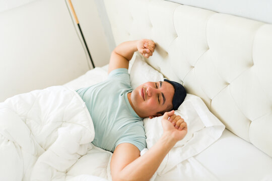 Latin man waking up in bed while stretching and feeling well rested after sleeping