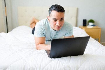 Young man comfortably works from home, typing on his laptop in bed, utilizing modern technology for his freelance work