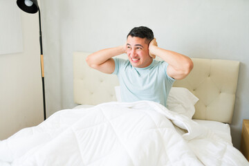 Annoyed young man sitting on a bed and covering his ears to block out loud noise