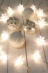 White with silver Christmas balls lie on a white wooden table along with a luminous garland. View from above.