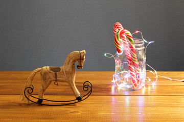 Christmas decoration - small wooden toy rocking-horse and caramel New Year's candies in a glass jar entwined with a luminous festive garland on a table against a dark blue wall.
