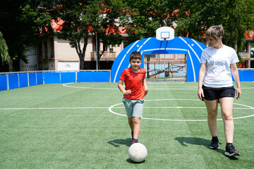 Young boy and his mother walking on a sunny soccer field, the boy kicking the ball forward.
