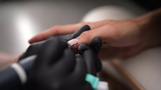 Close-up of unrecognizable beautician wearing black gloves using electric manicure drill to remove cuticles from male client finger at nail salon. Concept of importance of nail care and self-care.