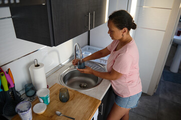 Woman in casual outfit washing dishes at the kitchen sink with natural light streaming in through the window