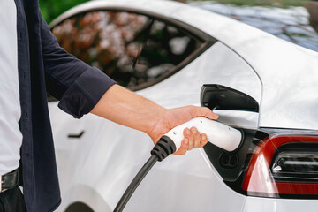 Man recharging battery for electric car during autumnal road trip travel EV car in autumnal forest. Eco friendly travel on vacation during autumn with electric vehicle. Exalt