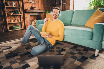 Obraz premium Full body photo of nice young man sit floor watch video netbook eat chips wear yellow clothes modern interior flat indoors
