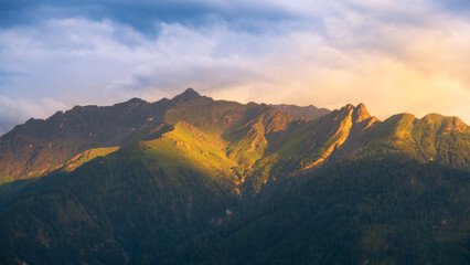 Majestic mountain peaks glow with warm golden light as the sun sets, casting long shadows and creating a serene atmosphere in the sky.