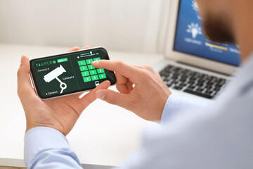 Young man using mobile phone with smart home security system application at table in room, closeup