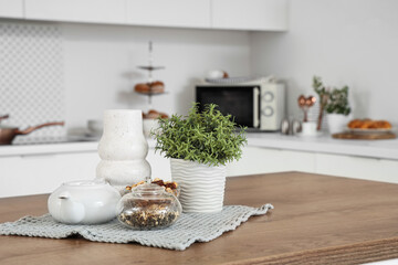 Teapot with bowl of dried tea and plant on table in kitchen, closeup