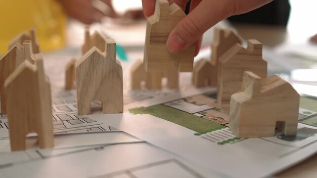 Architect and engineer discuss about building blueprint on table with architectural document, measurement tools scatter around at office, use wooden block for house positioning. Closeup. Delineation.