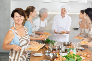 Elderly woman in apron learns to cook chicken dish at cooking master class