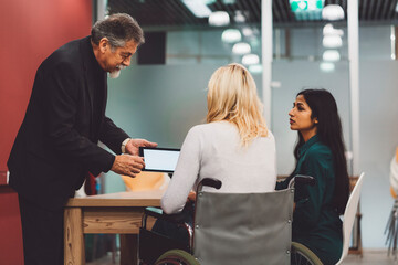 Obraz premium Senior CEO holding a tablet showing something to a disabled blonde woman and young Indian woman