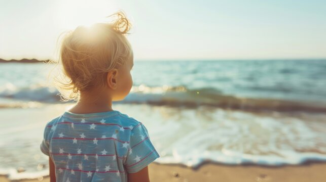 A little girl stands on the beach, watching the waves as she proudly displays an American flag in the warm sunlight