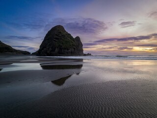Sandy beach and rocky cliff at low tide with pools and reflections in the sand at sunset.  Piha,...