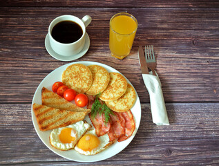 Continental breakfast, scrambled eggs, crispy bacon, cherry tomato croutons and pancakes on a wooden table, next to a cup of hot coffee and a glass of orange juice.