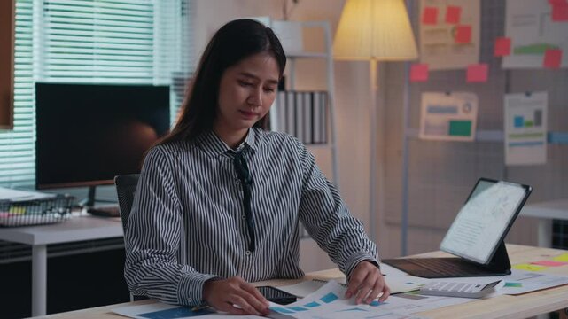 Young businesswoman is reviewing financial data and reports at her desk in a modern office. She is using a tablet and has a variety of documents spread out in front of her