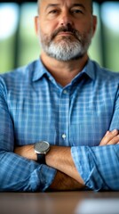 Fototapeta premium Confident man with beard and blue shirt poses with arms crossed, showcasing professionalism and authority in a modern workspace.
