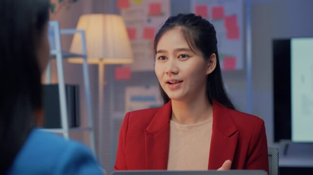 Young asian professional woman in a red blazer smiles while engaging in conversation with a recruiter in a modern office setting, showcasing confidence and positivity during the job interview