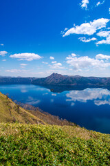 雲の浮かぶ青空を反射する山間の湖。日本の北海道の摩周湖は美しい自然の景色が観光お客に人気の名所。雄大で迫力のあるカルデラ湖の風景が魅力。