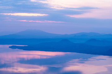 薄紫色の幻想的な夜明けの空を反射する湖と周囲の山々のシルエット。少し霞んだ印象の穏やかな自然の景色。