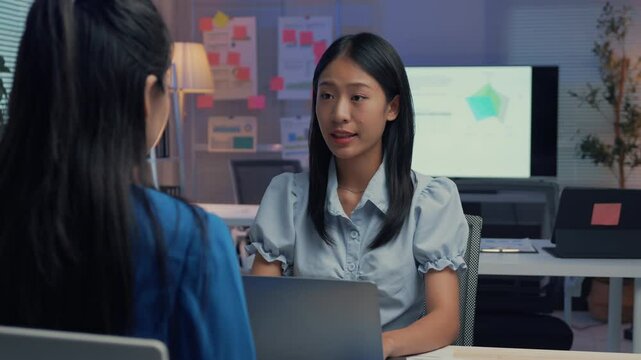 Asian manager explains concepts to a colleague in a modern office at night. The scene highlights teamwork and effective communication in a corporate setting