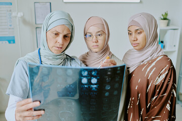Medical professional analyzing X-ray while two patients wearing hijabs attentively observing the results in a clinic setting