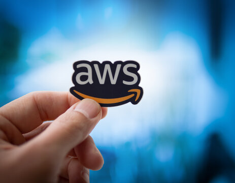 LOS ANGELES, CA - June 1, 2024: Human hand holding Amazon Web Services AWS logo sticker in front of glowing blurred computer screen background. Closeup.