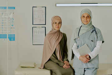 Obraz premium Portrait of female doctor standing beside seated patient, both wearing hijabs, in clinical setting with medical charts and equipment visible in background