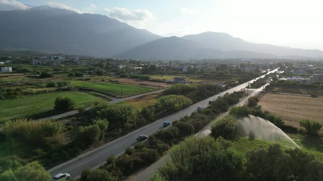 The drone flies over a busy road with visible cars, showing field irrigation in the foreground and towering mountains in the distance, with the sun shining through light white clouds.