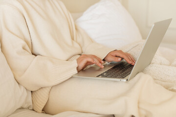 Fototapeta premium Close-up of a young woman in a cozy, light-colored house outfit working on a laptop at home in bed.