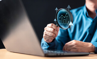 financial, analysis, finance, investment, risk, strategy, marketing, report, research, search. A man is holding a magnifying glass over a laptop with graph on it. The graph is showing a positive trend