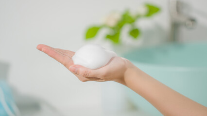 Close up shot at a cluster of white foam of bathroom skincare product on hand model palm, against on blurred bathroom background. Front view photo, blank space for designing
