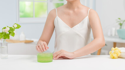 Woman in white dress sitting next to white table and preparing to apply lotion after shower. Mockup for advertising with a green unbranded cosmetic jar placed on table, space for product presentation