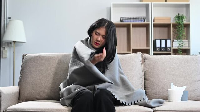 Young Asian woman suffering from flu symptoms, covered with a blanket on a sofa. Concept of illness, healthcare, and recovery.