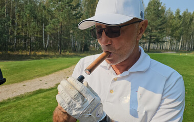 A golfer enjoys a cigar while relaxing on the course amidst green trees during a sunny day in early...