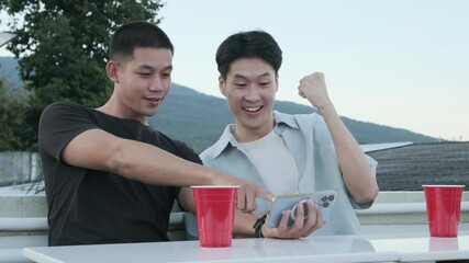 Two joyful asian male friends watching soccer game on mobile phone and celebrating victory  - Powered by Adobe