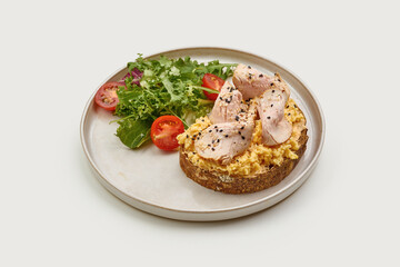 Breakfast plate featuring a slice of whole-grain toast topped with scrambled eggs, smoked turkey, and sesame seeds. A fresh side salad of cherry tomatoes and greens adds a vibrant touch.