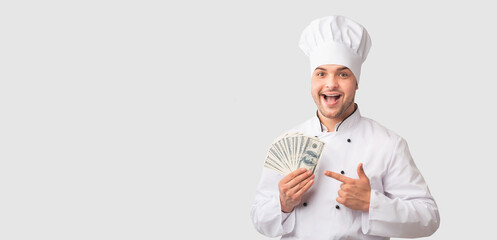 Restaurant Business. Excited Chef Man Holding Money Pointing Finger At Cash In His Hands Over White Background. Panorama