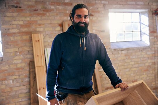 Smiling carpenter with dark beard, standing in his workshop and working with wood. He looks directly into the camera.