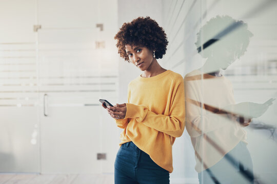 Young black businesswoman leaning against a glass wall and texting on her phone. She looks into the camera smiling.