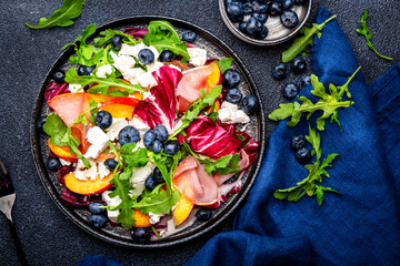 Foodie food salad with peach, jamon, soft cheese, blueberries and arugula on black background, top view