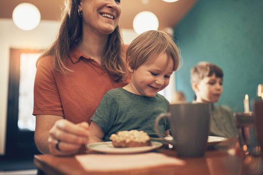 Adorable little boy smiling while sitting on his mother's lap next to his brother and having a snack during a family arts and crafts class at the weekend