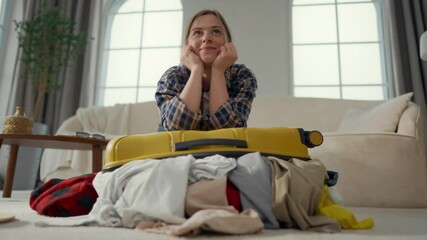 Portrait of dreaming female tourist with overload unclosed suitcase at home. Dream about holidays and trip in summer vacation, young student girl packing bag in living room, waiting for adventure